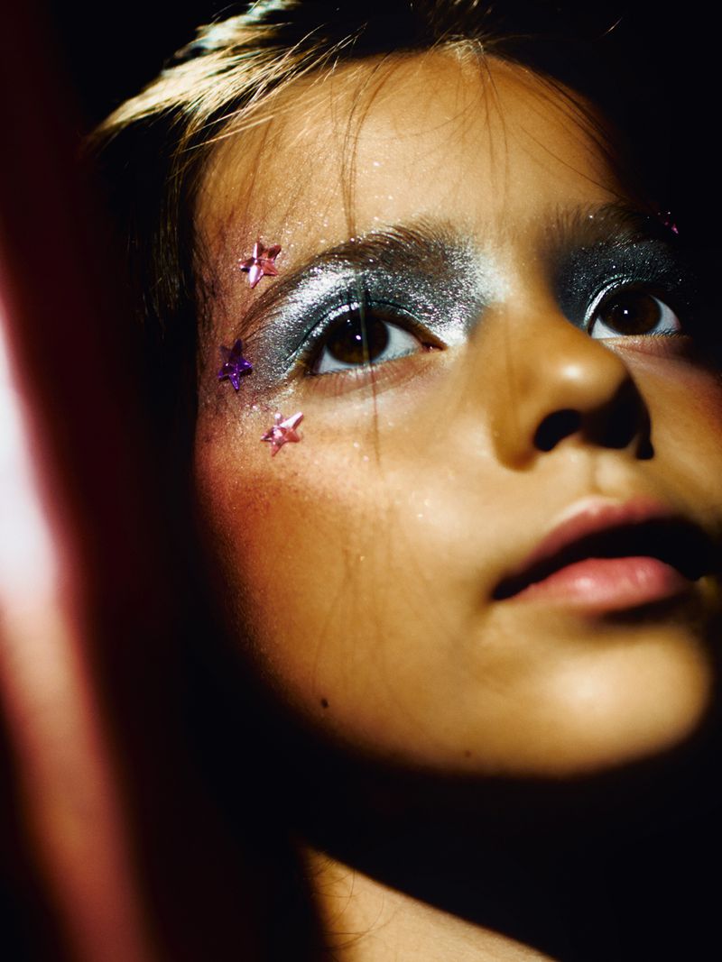 Close-up portrait of a young person with creative silver eyeshadow and star-shaped face jewels in front of a dark, shadowed background.