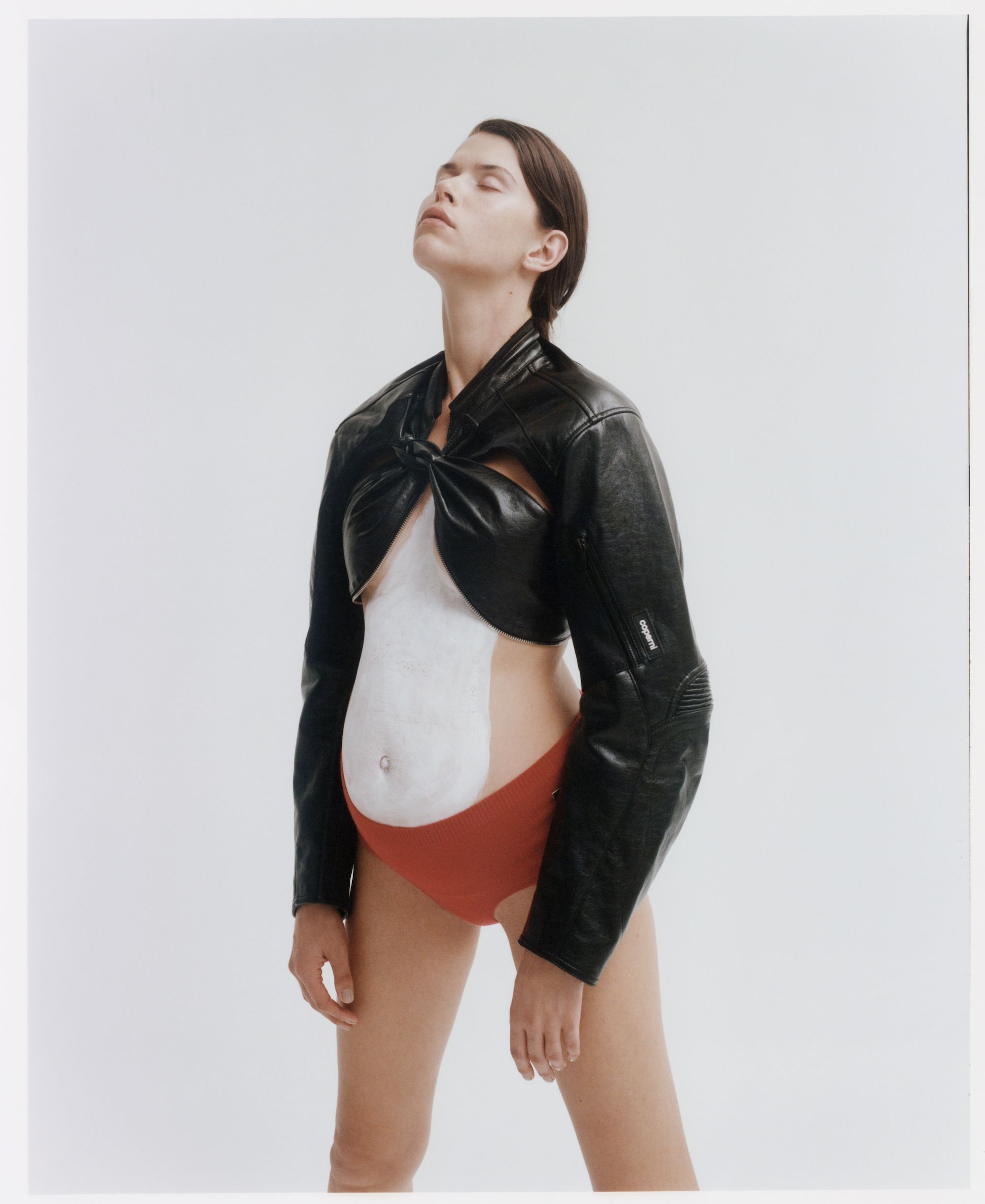 Studio shot of a woman with body paint wearing a short black leather jacket and red underwear in front of a white background with eyes closed.