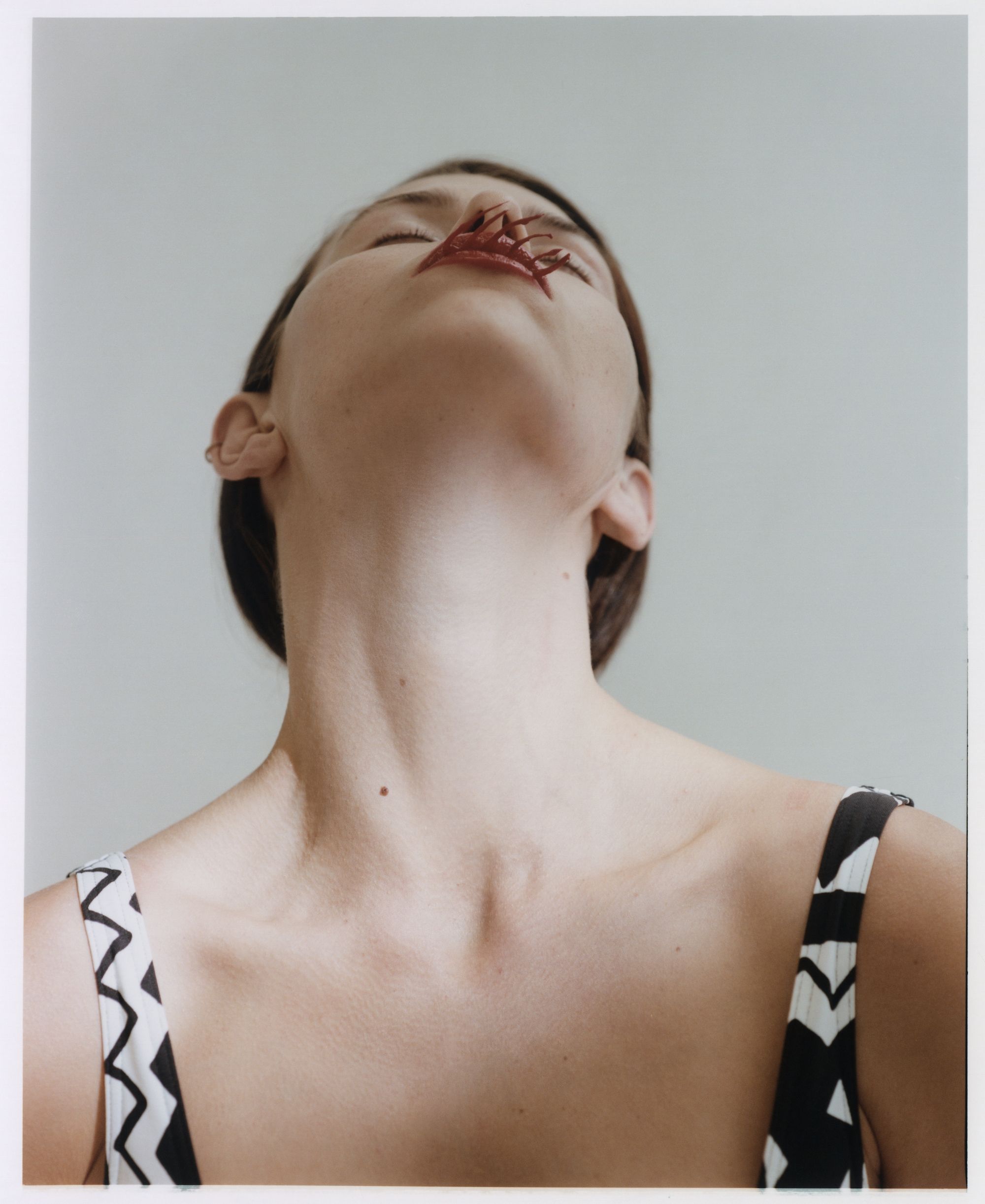 Low angle view of a woman with closed eyes and a flower on her face, wearing a patterned top against a light grey background.