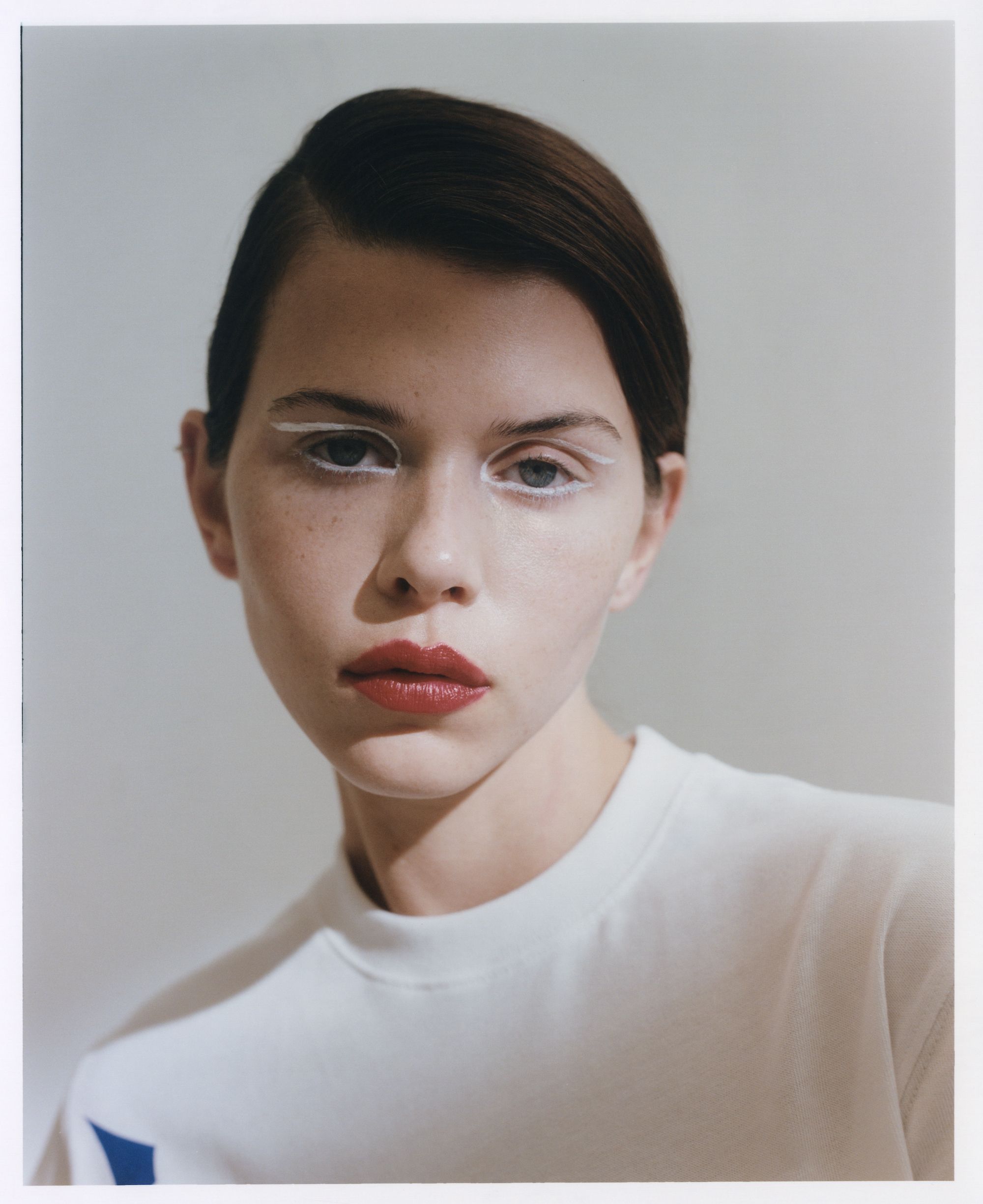 Close-up portrait of a young adult woman with white eyeliner and red lipstick, wearing a white shirt against a neutral background.