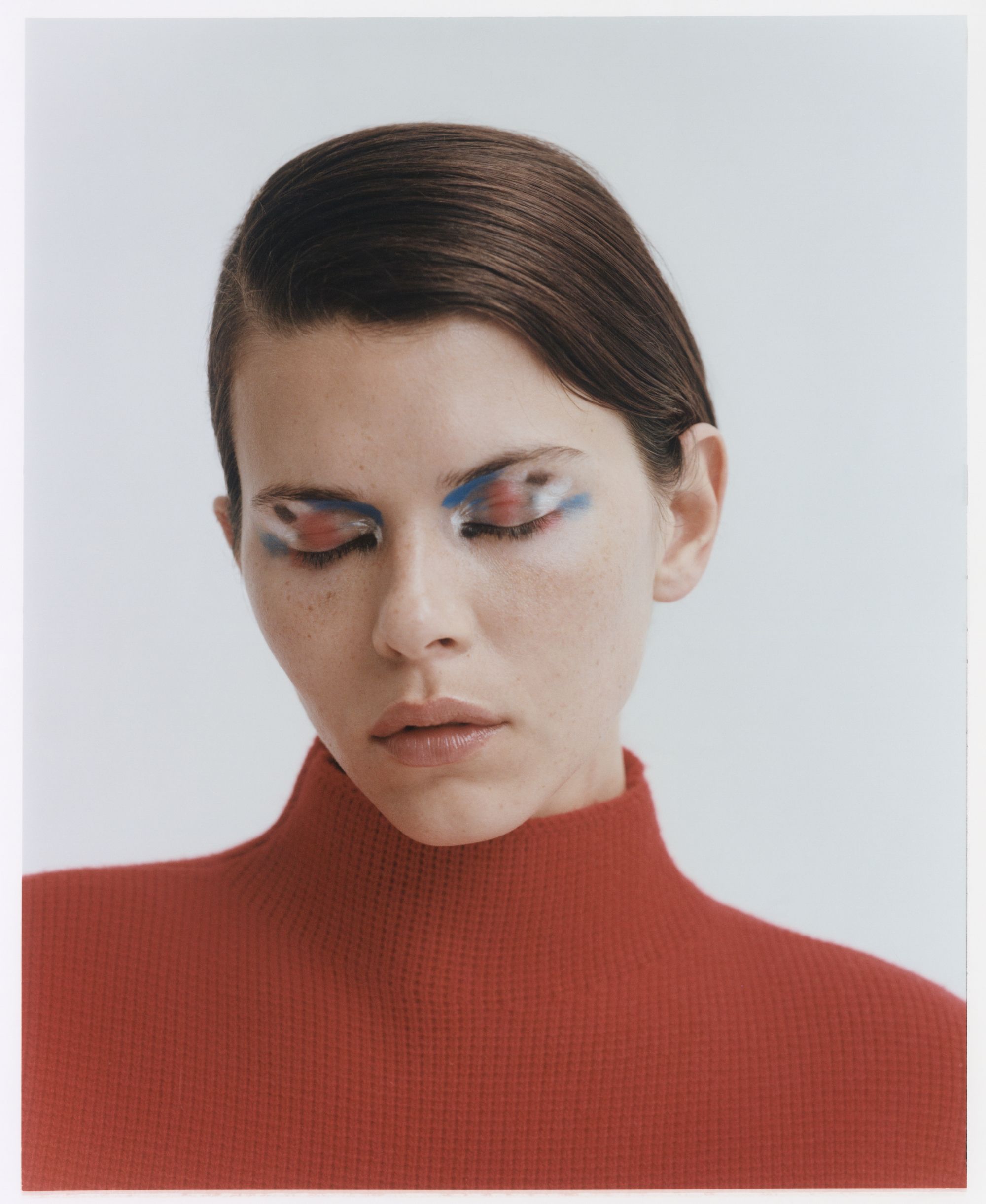 Close-up studio portrait of a woman with dramatic makeup and a red turtleneck sweater against a plain white backdrop.