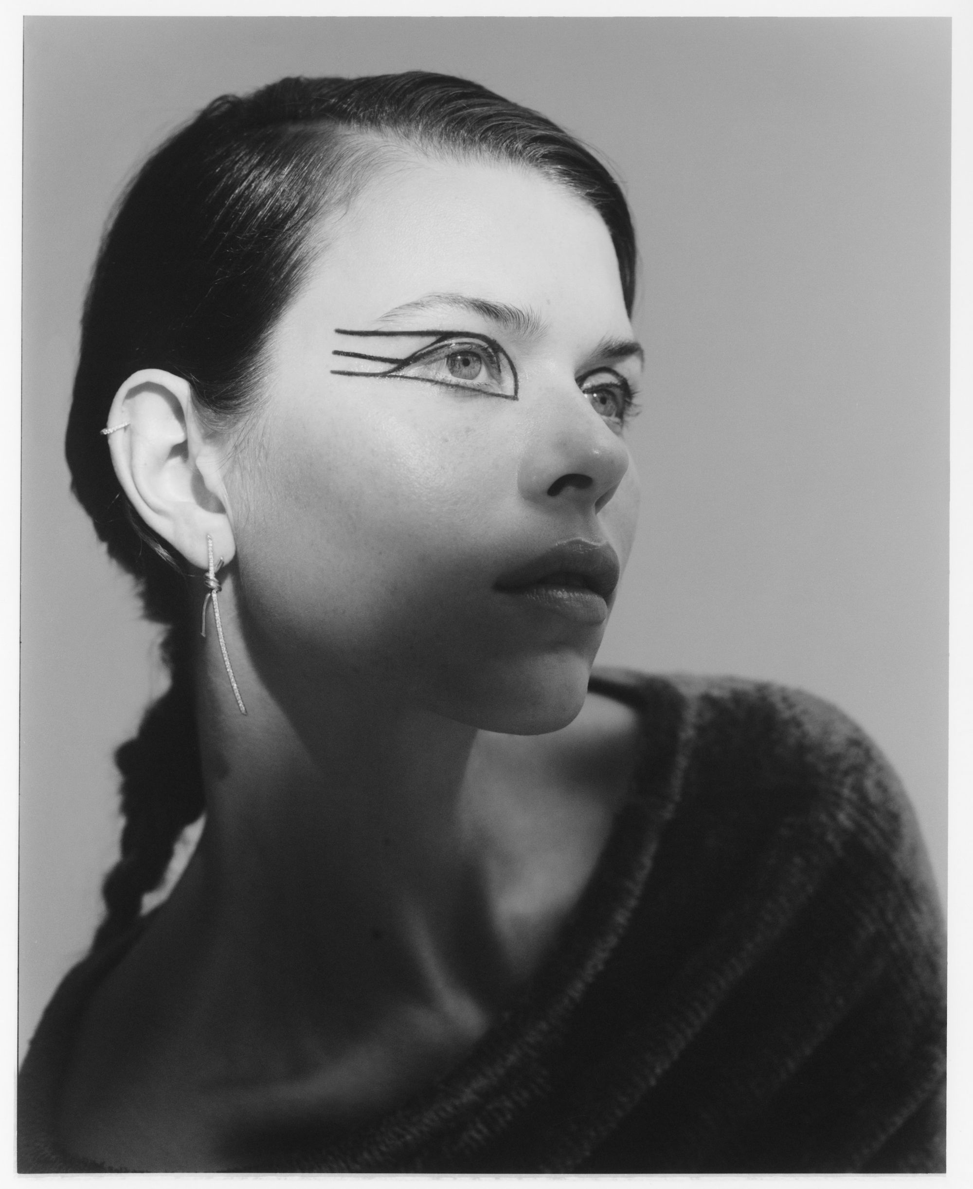Black and white portrait of a young woman with graphic eyeliner and a braided hairstyle against a plain gray studio backdrop.