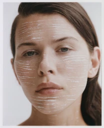 Close-up portrait of a woman with white cosmetic marks streaked across her face against a plain background