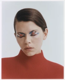 Close-up portrait of a woman with slicked-back brown hair wearing a red turtleneck sweater against a white seamless background.