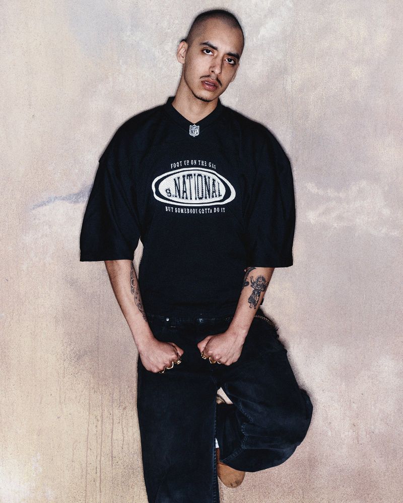 Model wearing a black t-shirt and dark jeans leaning against a textured neutral background for Willy Chavarria.
