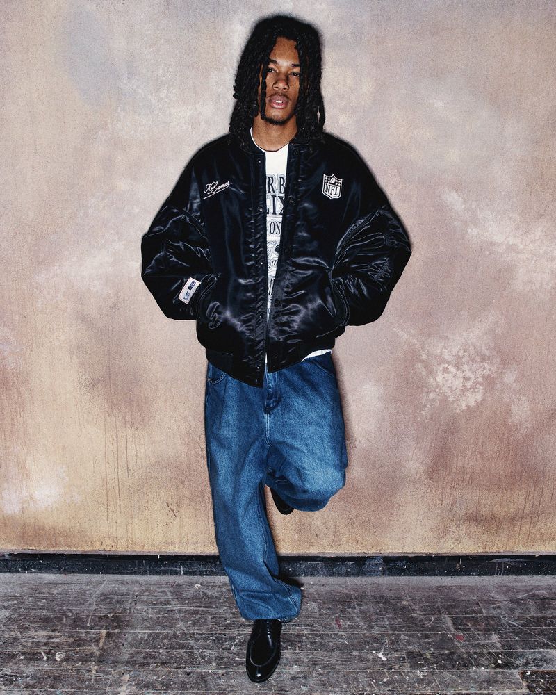 Young adult with dreadlocks posing in a black NFL bomber jacket and baggy jeans against a neutral mottled wall and wooden floor.