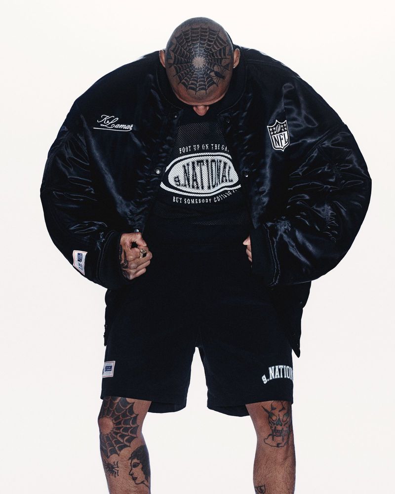 Adult male in a black jacket, shorts, and t-shirt stands indoors against a plain backdrop, showcasing distinctive hair tattoo.
