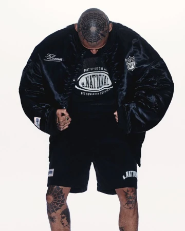 Adult male in a black jacket, shorts, and t-shirt stands indoors against a plain backdrop, showcasing distinctive hair tattoo.
