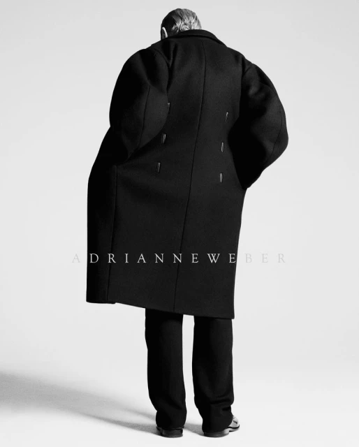 A person in a black coat and pants faces away from the viewer in a studio setting, focusing on fashion and editorial style.