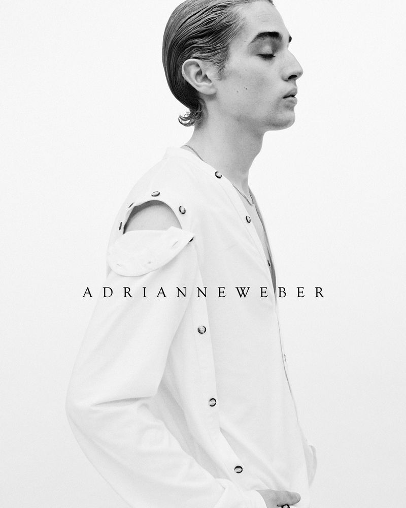 Monochrome portrait of a young adult with slicked-back hair, eyes closed, wearing a white shirt with metal grommets, against a white background.