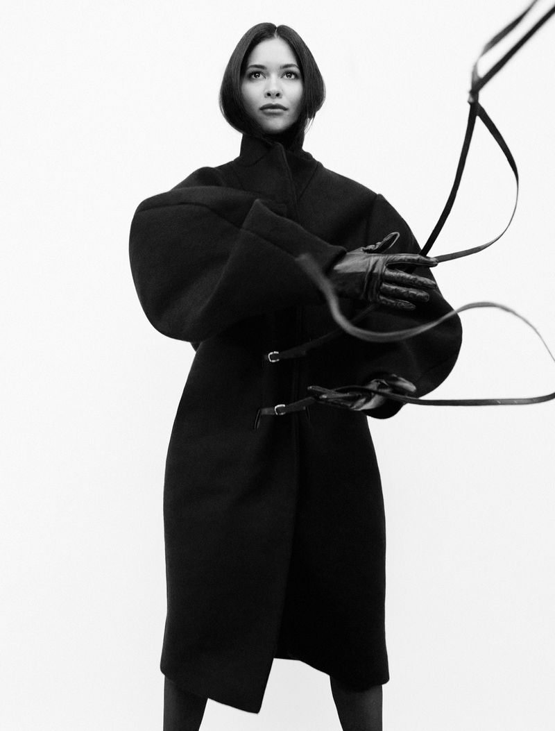 Fashion shot of a woman in a black coat and gloves, holding black straps against a white background in a studio, portrait orientation.