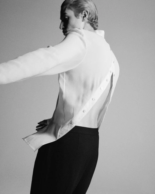 Elegant monochrome portrait of a slender man in a white shirt and dark pants, captured indoors with a minimalist grey background and studio lighting.