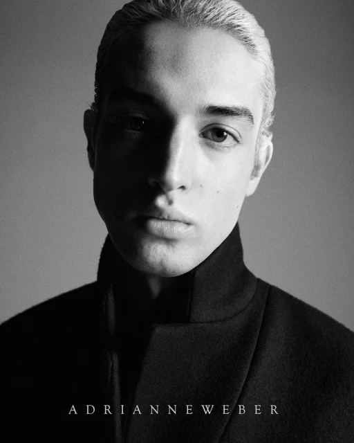 Portrait of a light-skinned young adult male with fair hair, wearing a black top with a high collar, shot in black and white.
