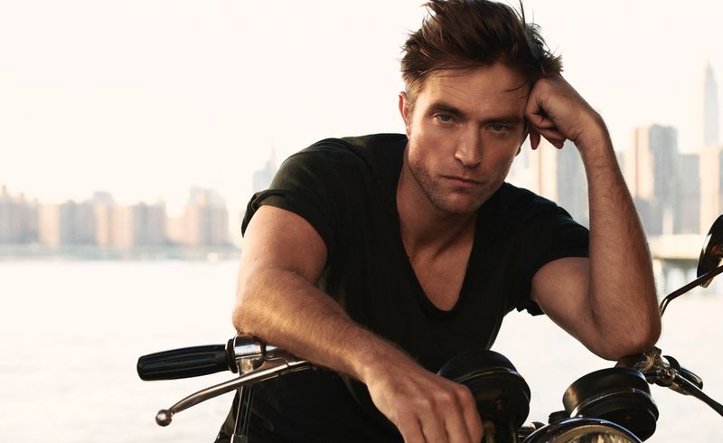 Portrait of a light-skinned adult male in a black shirt, posed on a motorcycle with a city skyline background, shot in bright daylight.