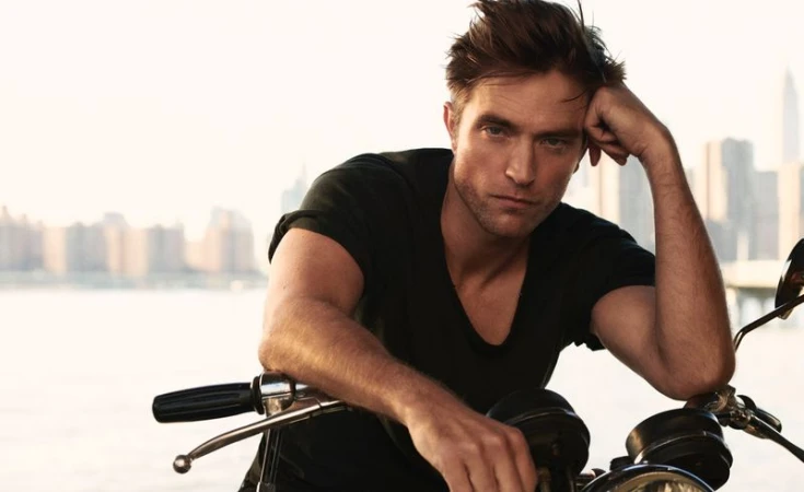 Portrait of a light-skinned adult male in a black shirt, posed on a motorcycle with a city skyline background, shot in bright daylight.