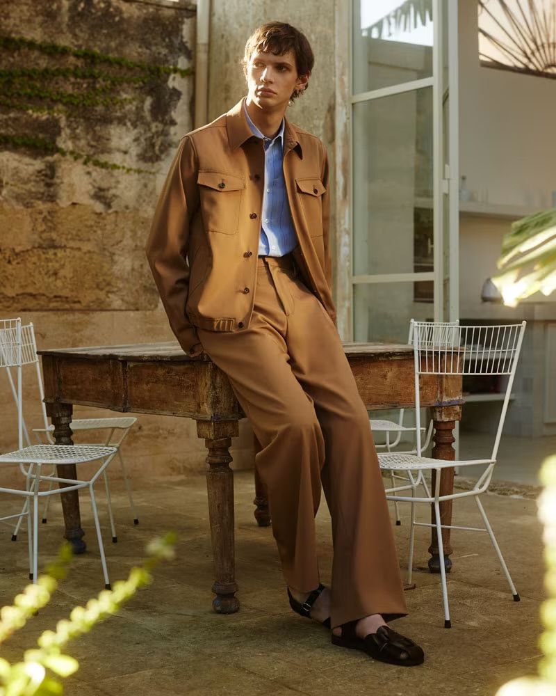 An adult male fashion model in a tan suit sits on an old wooden table outdoors with green vines on a stone wall in the background.