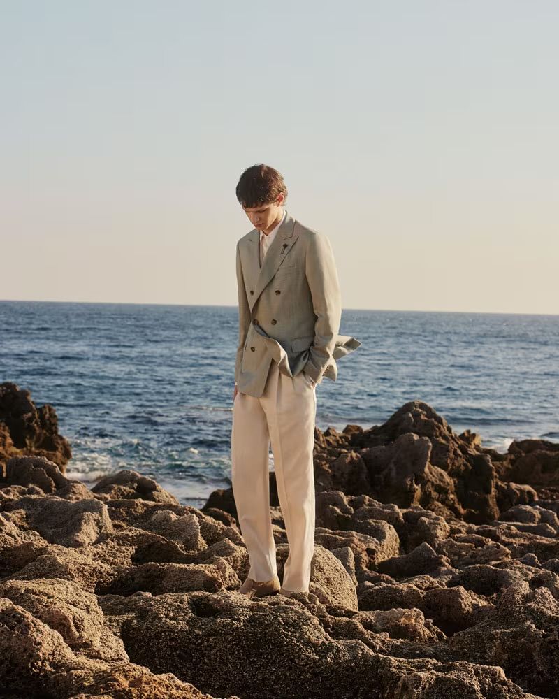 A person in a blazer and pants stands on a rocky shore by the ocean under a bright, clear sky, looking down at the rocks.