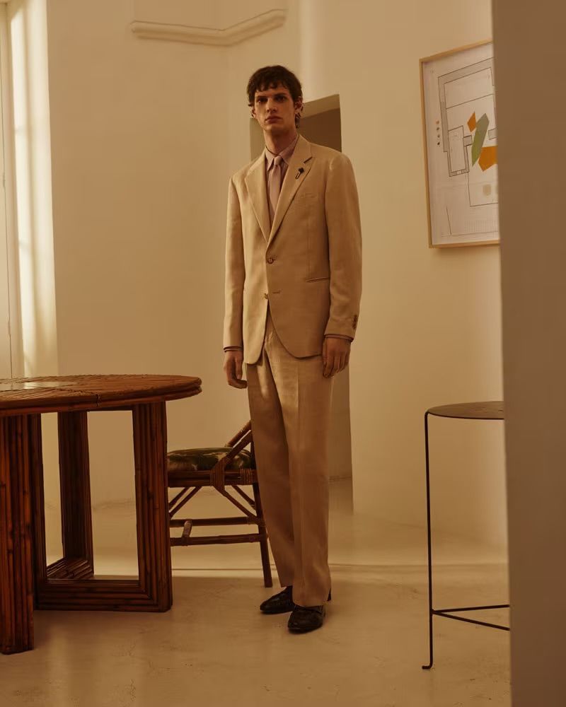 A young adult man stands in a light beige suit and dark shoes in an indoor setting with vintage-style furniture and an abstract art print.