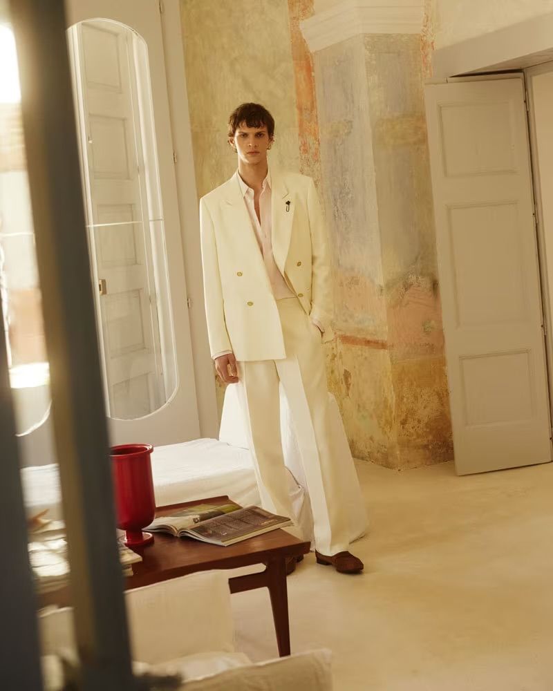 A stylish man in a double-breasted cream suit poses indoors in a fashion editorial shot with soft, diffused lighting and warm color grading.