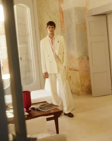A stylish man in a double-breasted cream suit poses indoors in a fashion editorial shot with soft, diffused lighting and warm color grading.