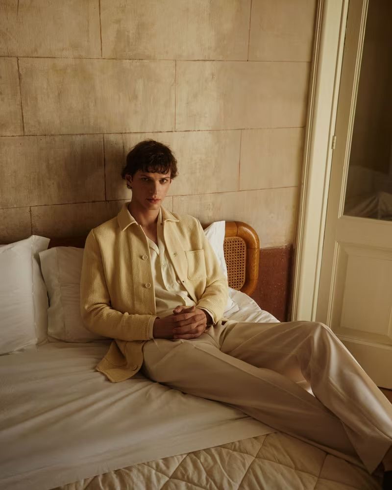 A tan-skinned man in a beige linen jacket and pants sits on a bed with white sheets against a neutral wall, bathed in warm, soft light.