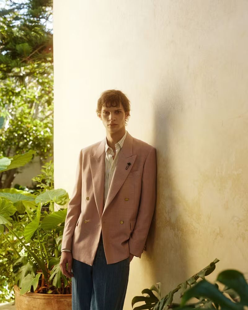 An adult male fashion model wearing a pink suit poses against a wall with green plants in the background in a lifestyle fashion shot.