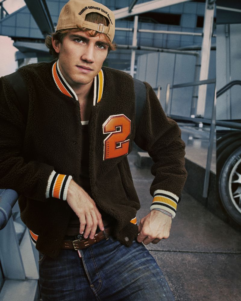 Attractive young adult male poses in casual fashion wearing jacket, jeans, and baseball cap; casual pose in urban setting.