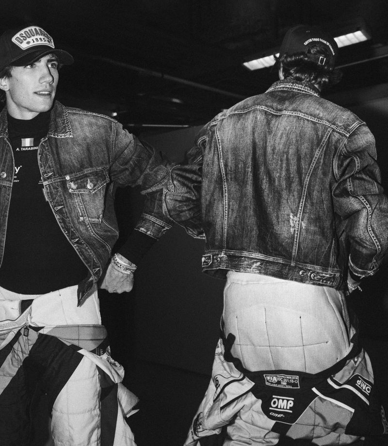 Black and white image of two adult males wearing racing suits and denim jackets standing indoors, with hats, one facing the viewer.