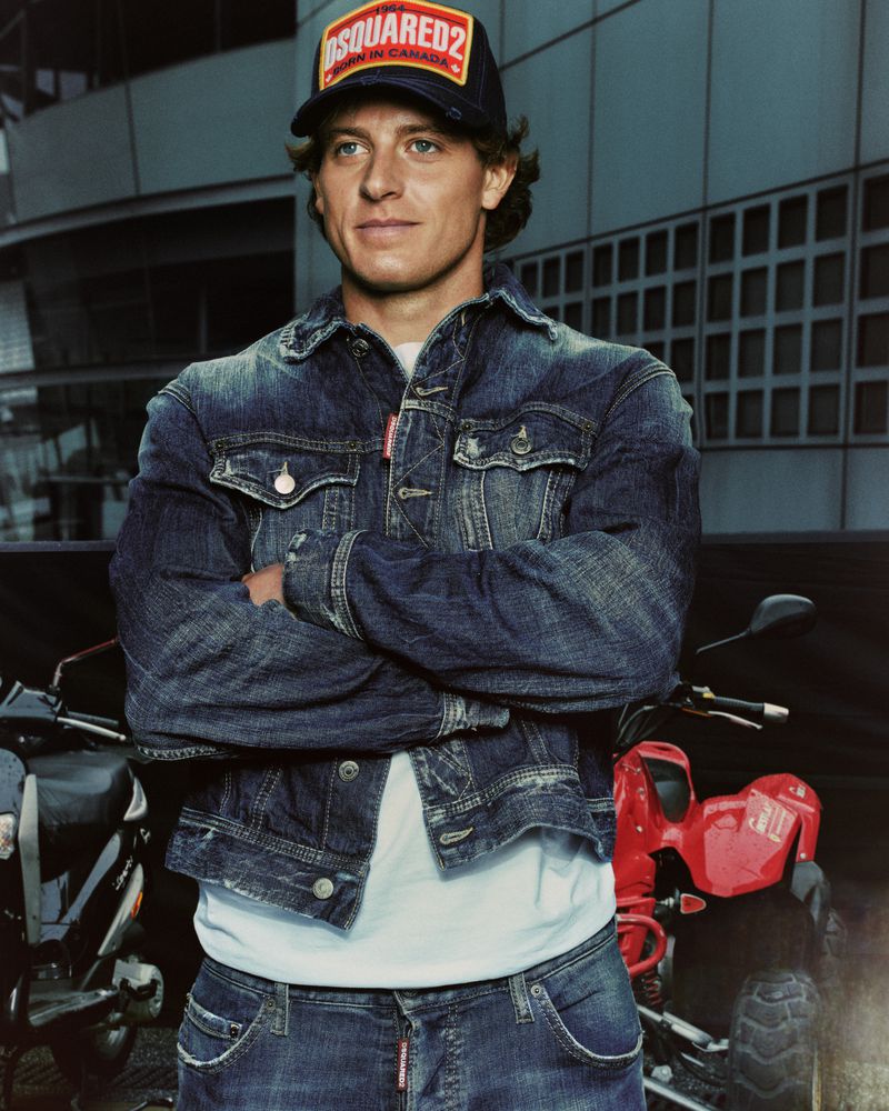 Man wearing a denim jacket and baseball cap standing with arms crossed outdoors with motorcycles
