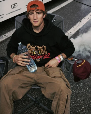 Young adult male sitting in a folding chair, holding water bottles, wearing a graphic hoodie, tan pants and an orange baseball cap.