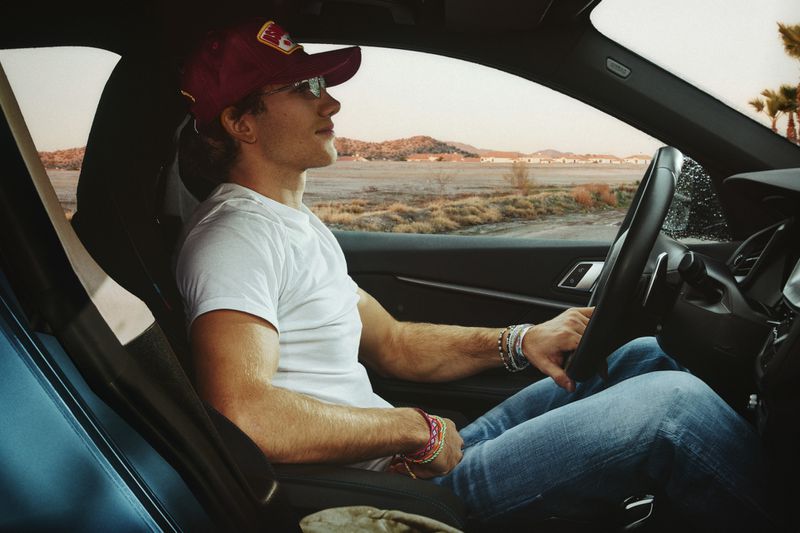 A man in a red baseball cap driving a modern car during the daytime, with a view of a desert-like landscape through the windshield.