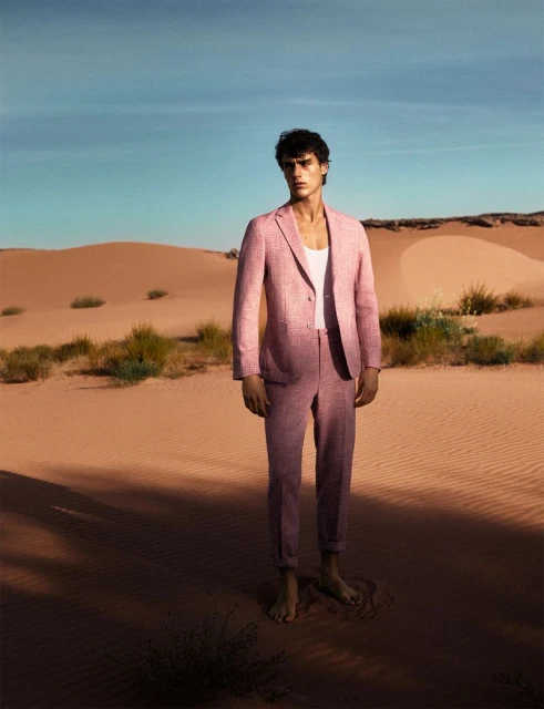 Man in pink suit standing in desert landscape with sand dunes, blue sky. Fashion editorial shot, outdoor setting, stylish male model.