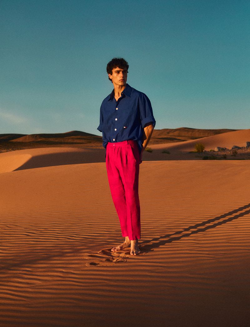A young adult man stands barefoot on a sand dune wearing a blue shirt and pink pants in a desert setting with a clear blue sky.