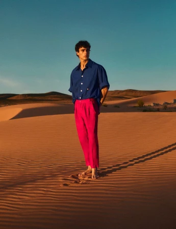 A young adult man stands barefoot on a sand dune wearing a blue shirt and pink pants in a desert setting with a clear blue sky.