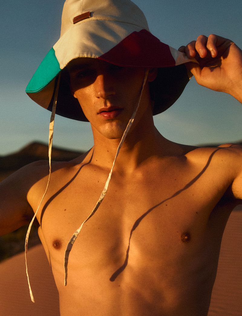 Portrait of a young adult male outdoors wearing a bucket hat with colorful panels, showcasing warm lighting and a sandy environment.