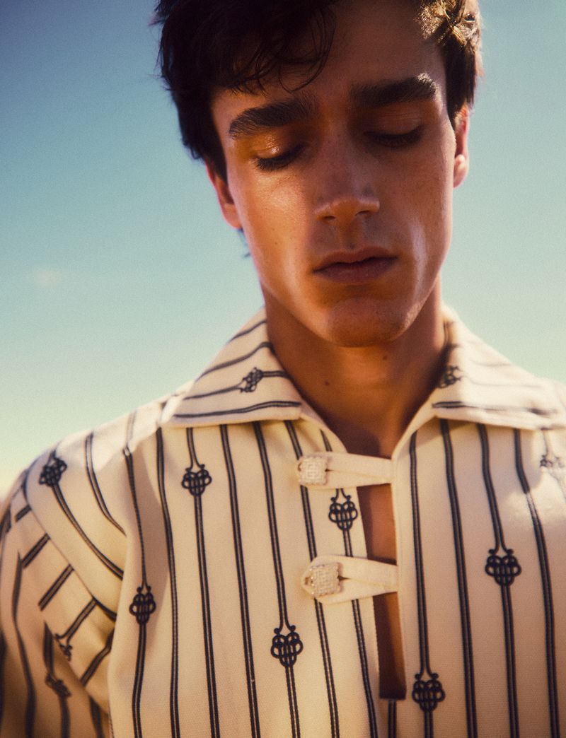 Close-up of a young adult male with dark hair and closed eyes, wearing a striped shirt against a clear blue sky in natural lighting.