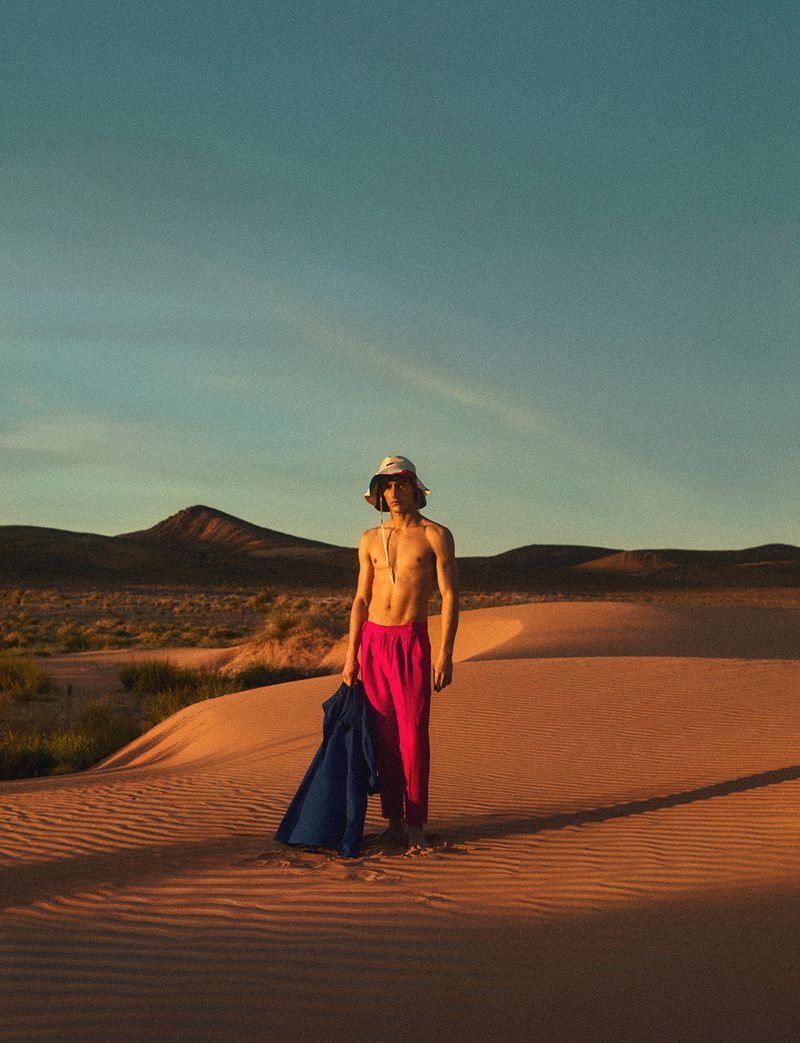 Young adult stands on sand dune with mountains and blue sky. Figure wears hot pink pants, no shirt, and bucket hat with jacket slung over arm.
