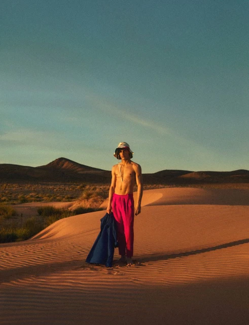 Young adult stands on sand dune with mountains and blue sky. Figure wears hot pink pants, no shirt, and bucket hat with jacket slung over arm.