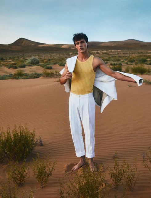 An adult male model in a yellow tank top and white pants stands barefoot in a desert landscape with a jacket draped over his shoulder.