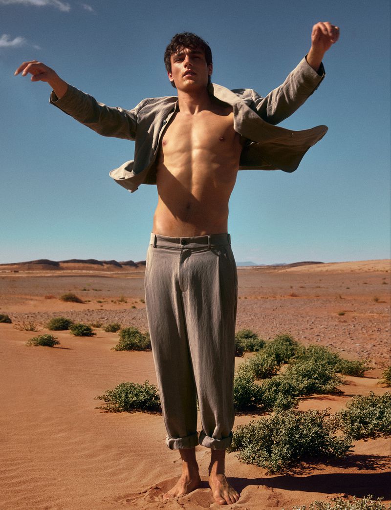Shirtless male model wearing grey trousers and an open jacket standing in a desert landscape under a clear sky.