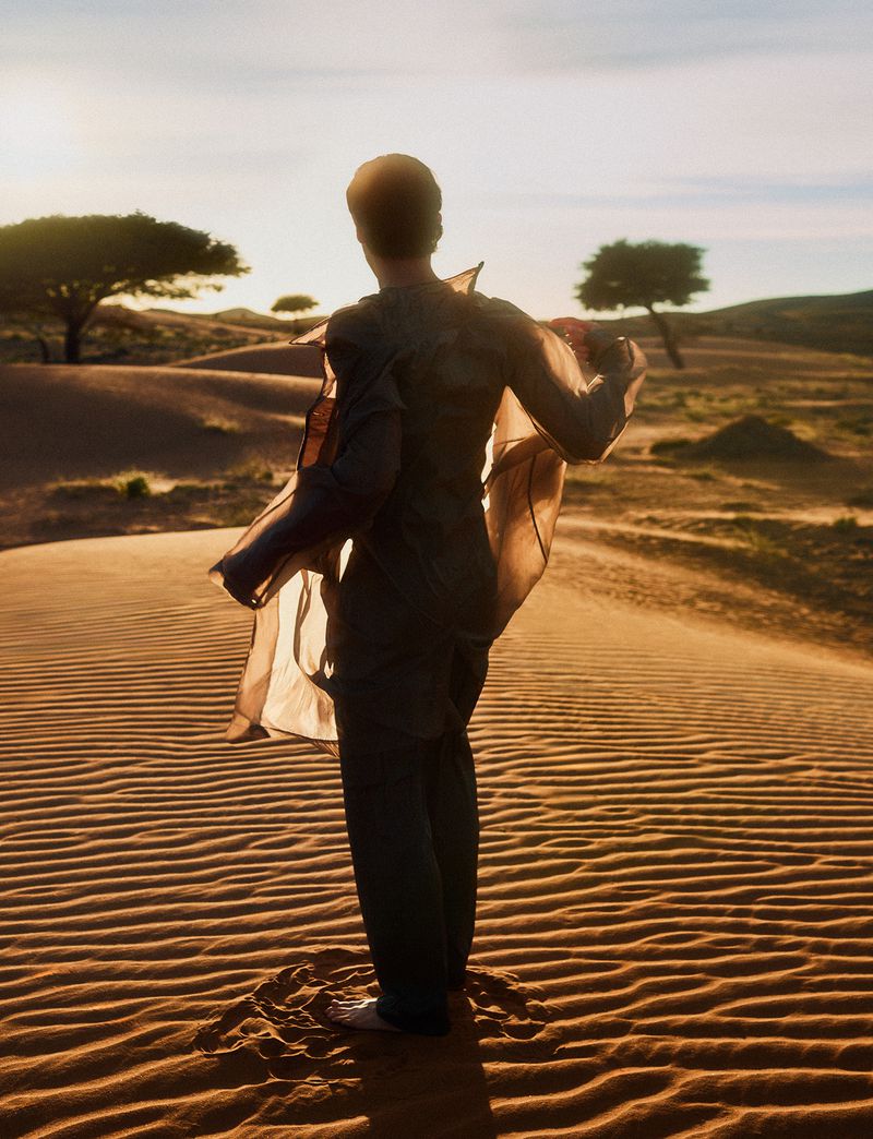 Back view of a person in desert with sheer fabric flowing in the wind, sand dunes and trees in the background, warm golden light.