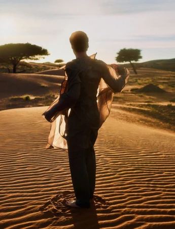 Back view of a person in desert with sheer fabric flowing in the wind, sand dunes and trees in the background, warm golden light.