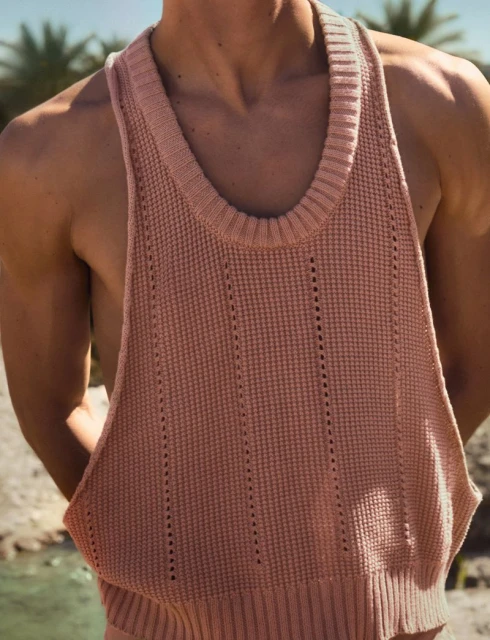 Close-up of a man with tan skin wearing a knitted pink tank top, against a background of water and palm trees on a sunny day.