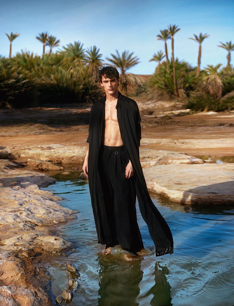 A male fashion model stands in shallow water in a black robe and pants in a desert environment with palm trees in the background.