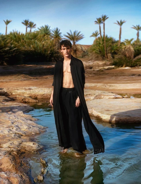 A male fashion model stands in shallow water in a black robe and pants in a desert environment with palm trees in the background.