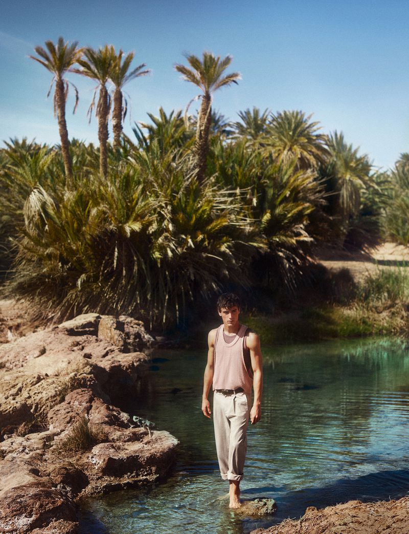Adult male wading in clear water, wearing a tank top and light pants, with palm trees and arid vegetation in the background under a clear sky.
