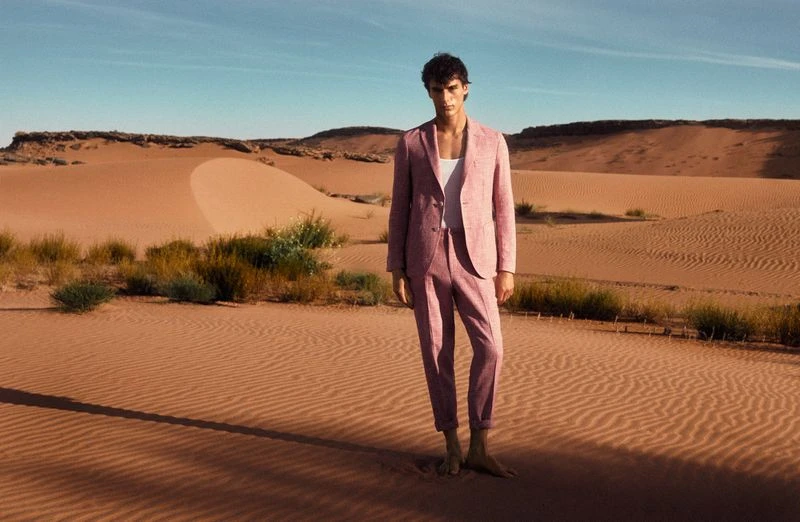 Male model in a pink suit stands barefoot in the desert against a backdrop of rolling sand dunes and a clear blue sky.