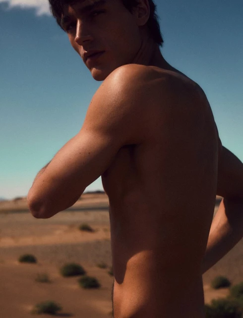 Torso shot of adult male with tanned skin and dark hair, outdoors, against a background of arid desert and a cloudless blue sky.