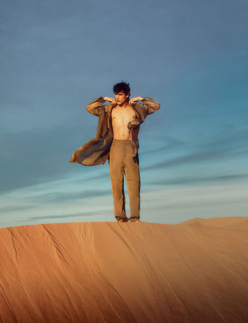 Man standing on a sand dune wearing an open jacket and trousers against a blue sky for Numero Netherlands Oasis editorial.