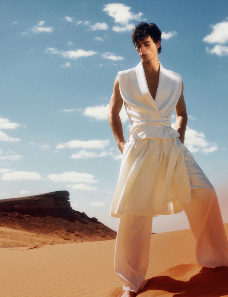 A fair-skinned man poses in a white sleeveless top and flowing white pants, set against a sandy desert landscape under a bright sky.